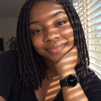 Jasmine with her chin on her hand, the Veyra on her wrist, by a window with blinds